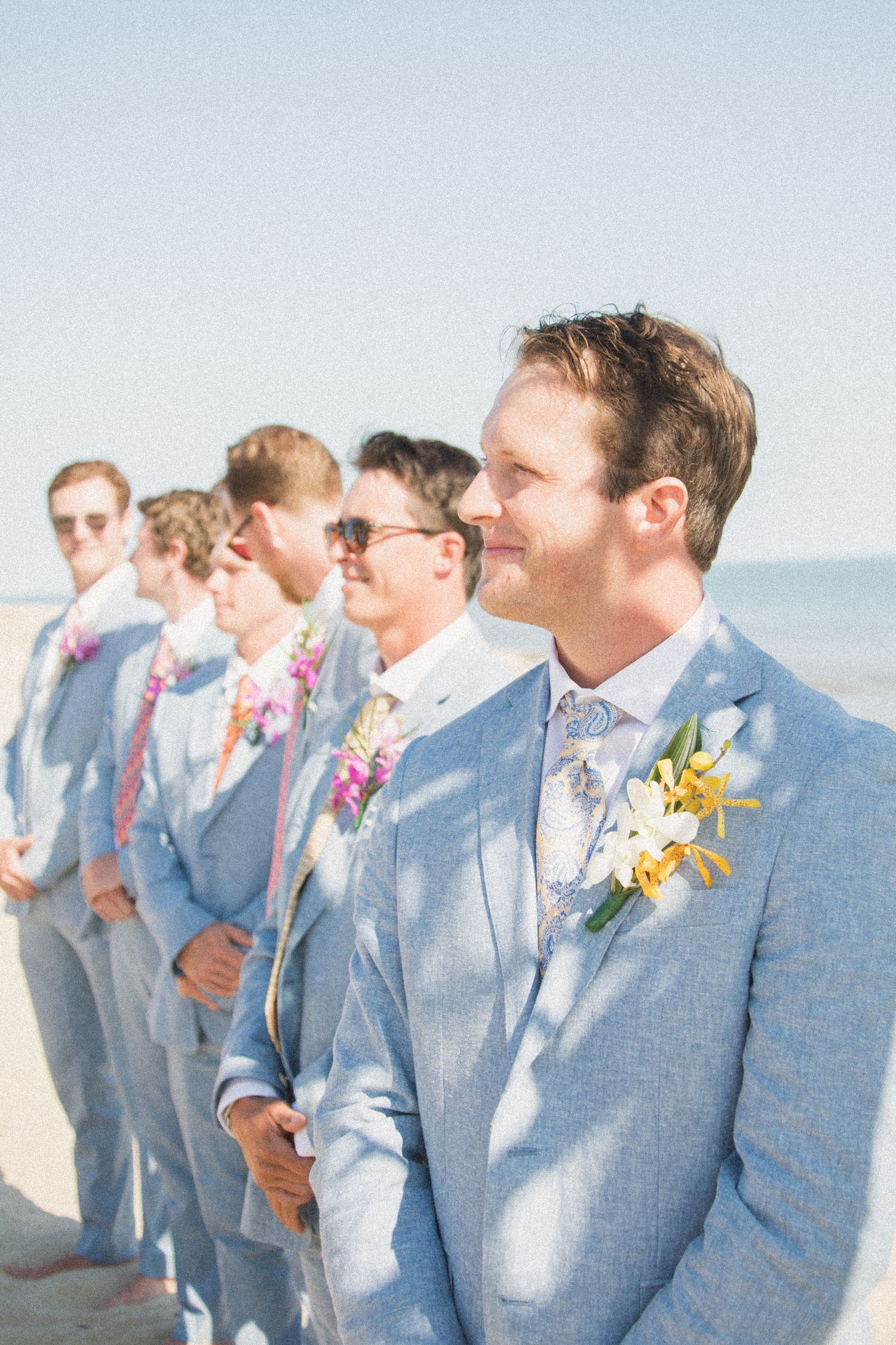 Wedding party in coordinated light blue suits and boutonnieres lined up on beach.