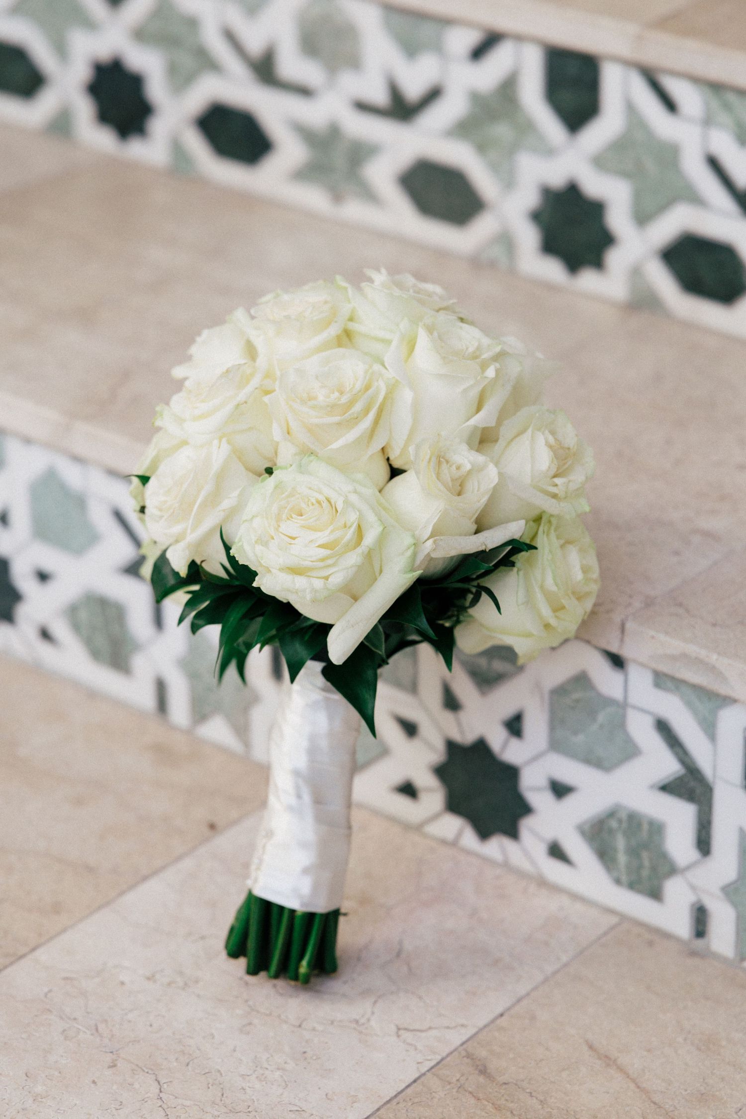 Elegant white rose bridal bouquet wrapped in silk ribbon against a geometric tile background.