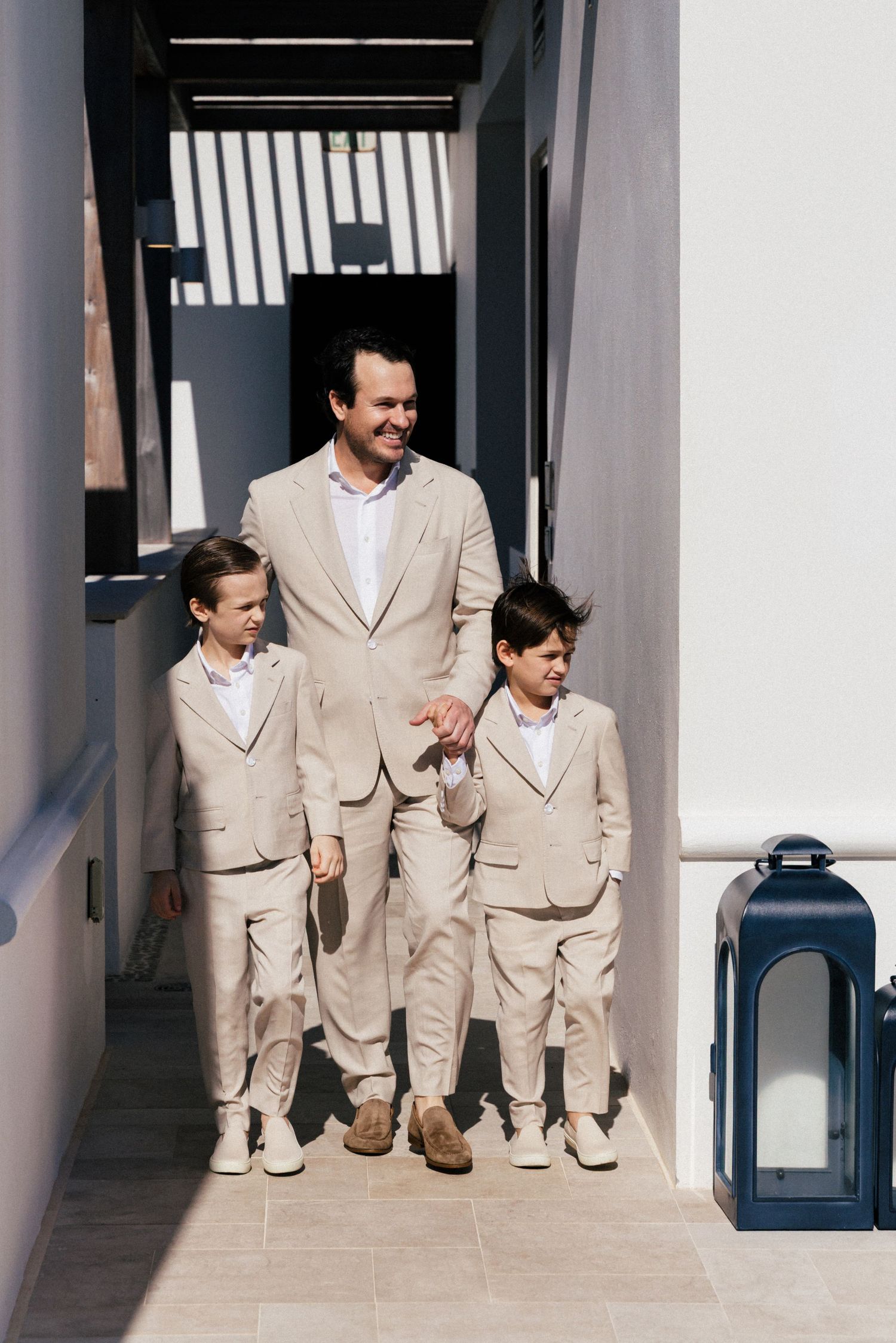 Three people in matching beige suits walk down a white hallway with lantern decor.