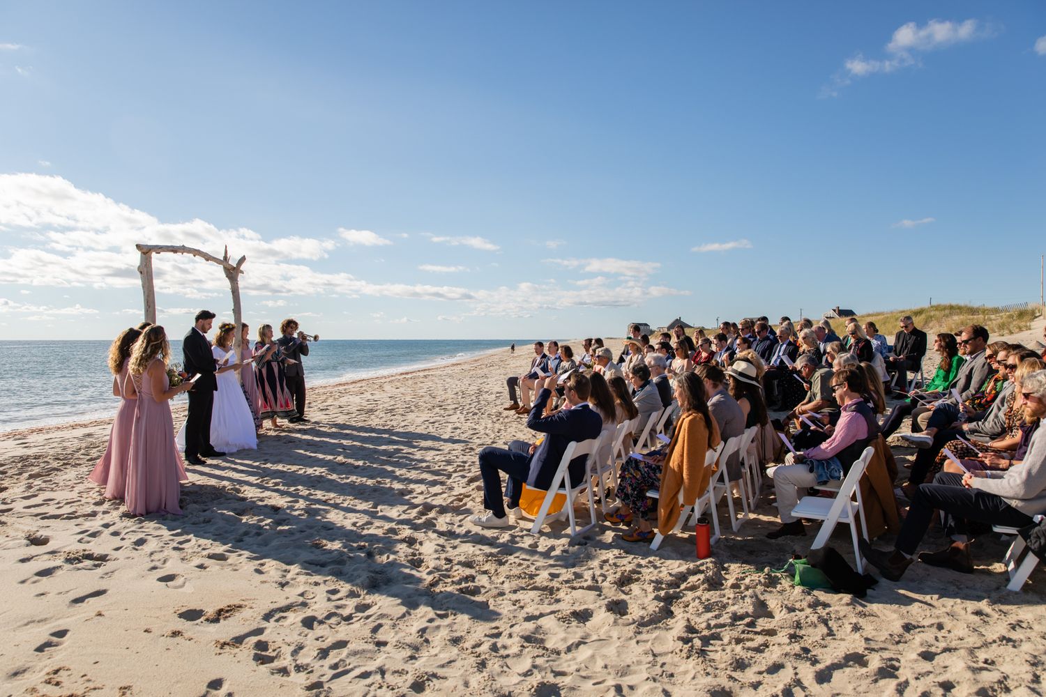 A beach wedding ceremony with guests seated in rows and an arch made of driftwood.