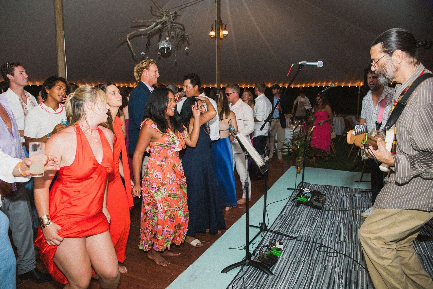 Large group of wedding guests dancing enthusiastically under a tent with string lights.