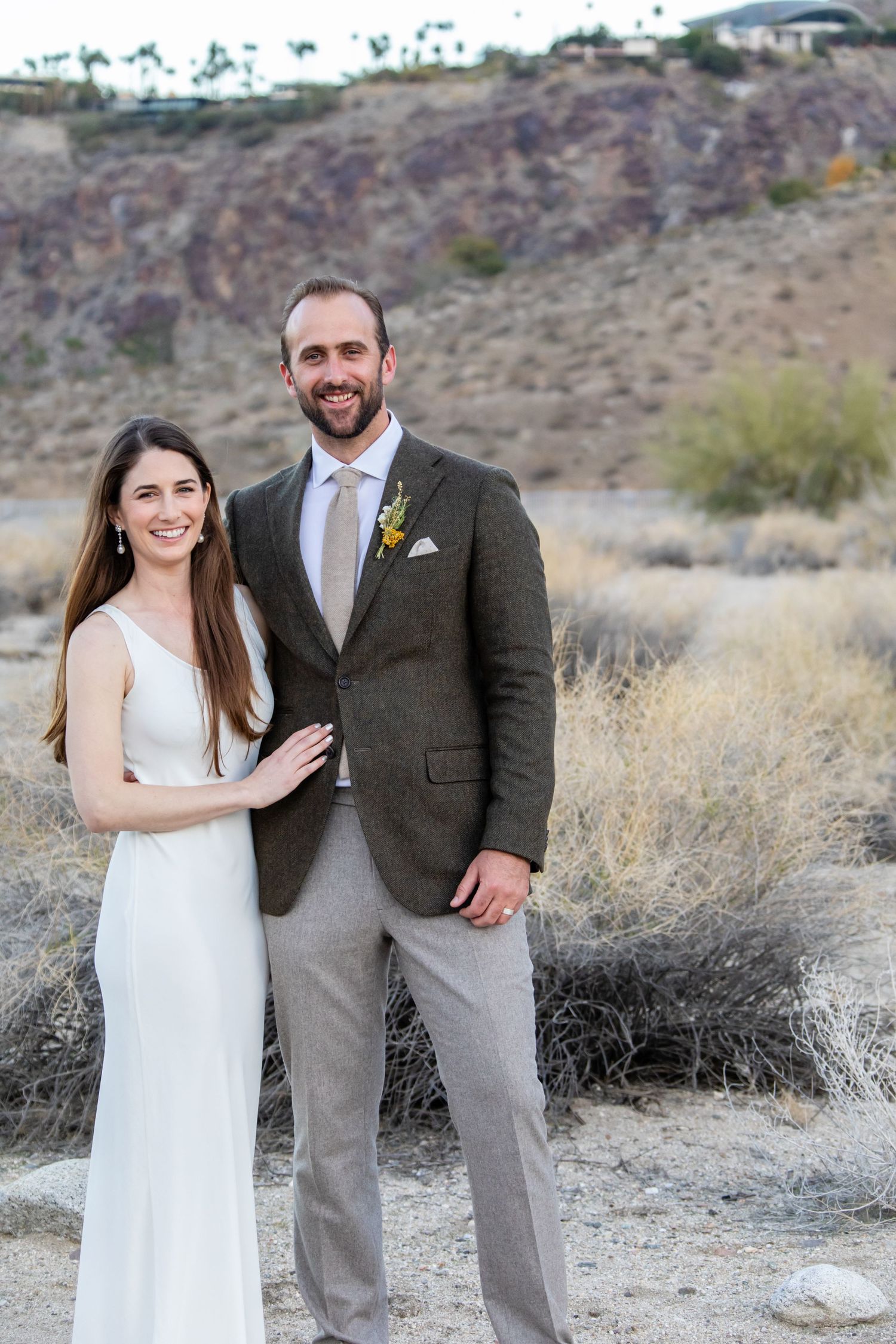 A couple poses for wedding photos in the desert with mountains and brush in the background.
