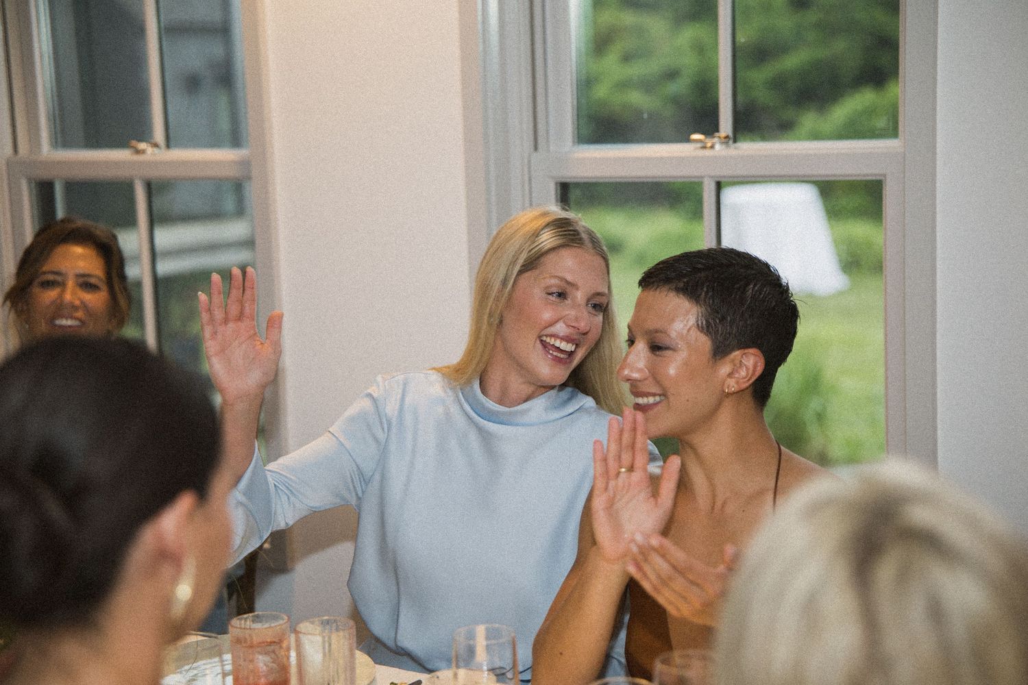 Women laughing and clapping together at a dining table near bright windows during a cheerful indoor gathering.
