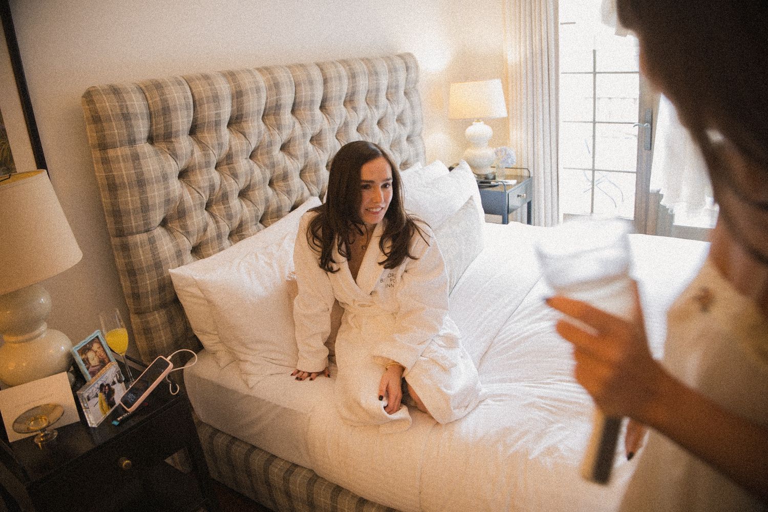 Smiling woman in a white robe sits on a tufted bed in a cozy bedroom while another woman holds a glass in the foreground.
