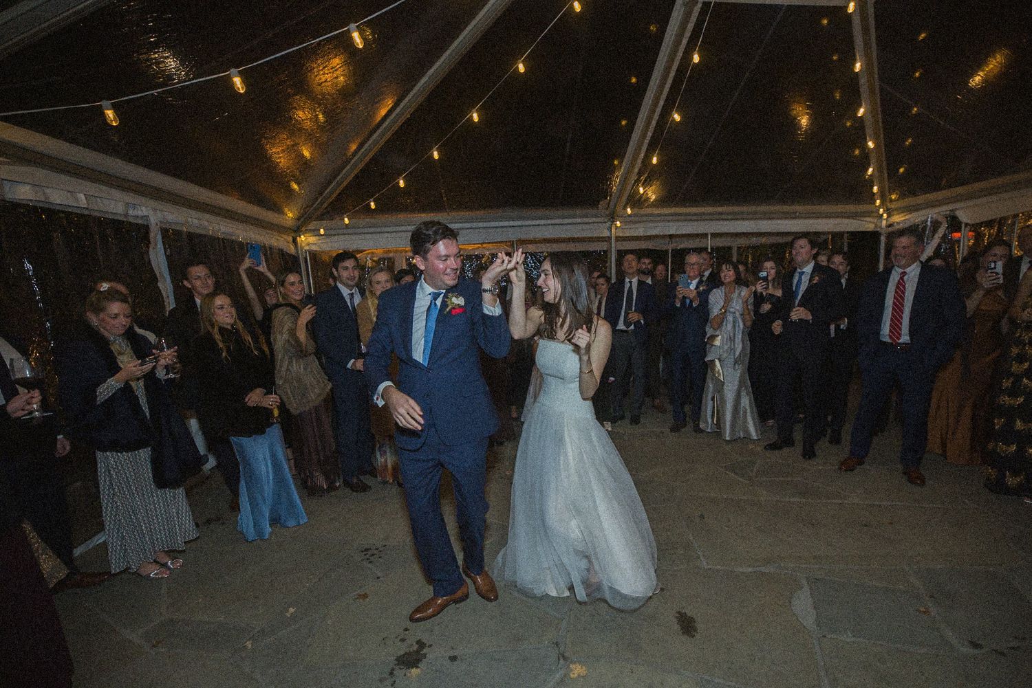 Bride in a white gown and groom in a navy suit share a first dance under a string-lit tent as wedding guests watch and applaud.