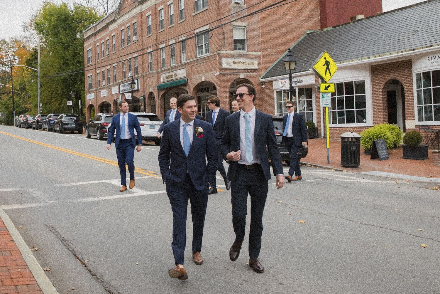 Two men in dark suits walk down a town street lined with brick buildings, with other groomsmen following behind.