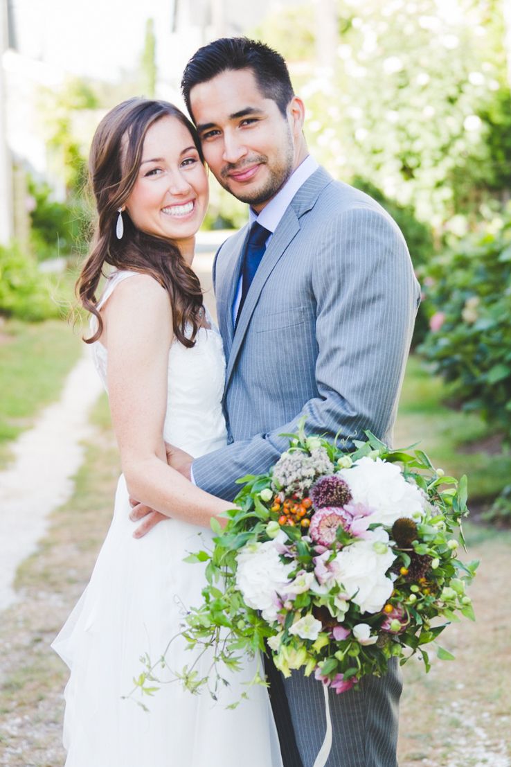 A couple poses with a vibrant floral bouquet in an outdoor garden setting.