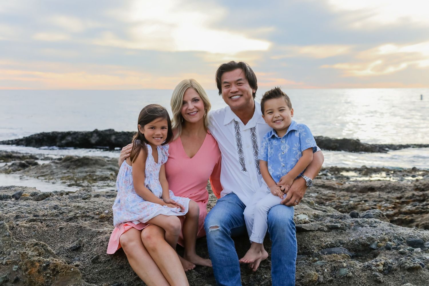 A family sits together on coastal rocks at sunset with the ocean in the background.