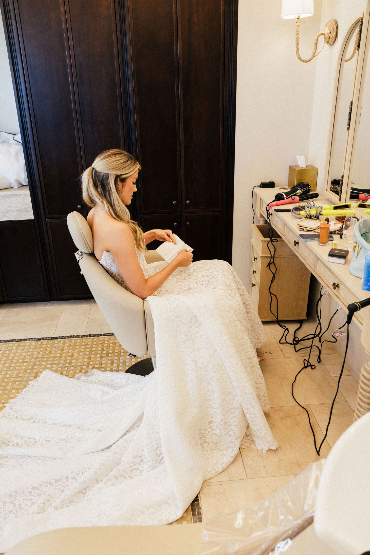 Bride in white lace gown getting ready at vanity mirror.