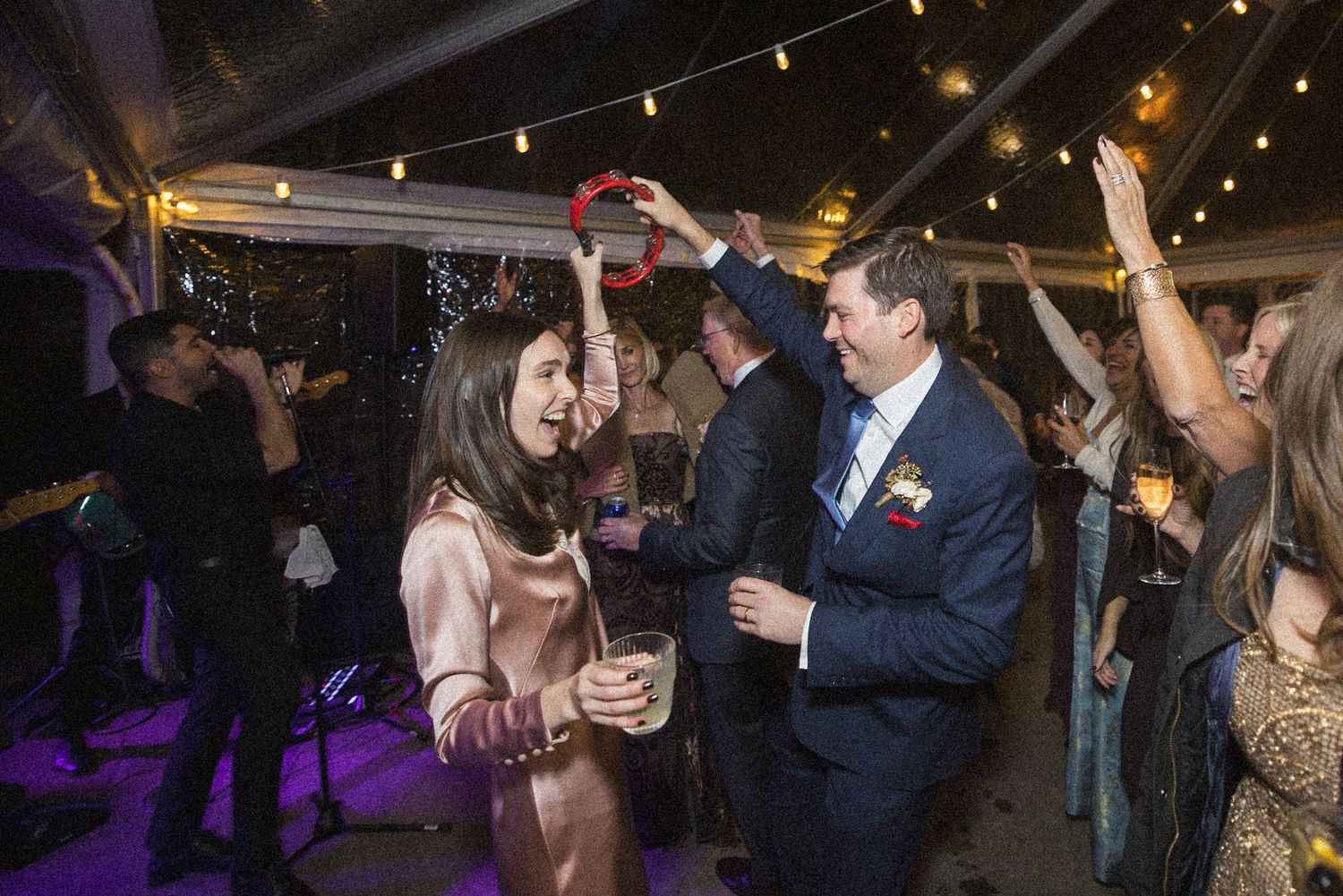 Wedding guests dance and cheer on a purple-lit dance floor under string lights, with a man in a navy suit raising his arm.