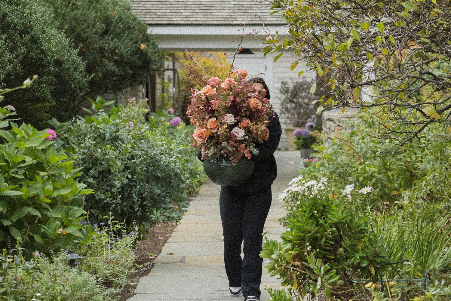 Person in dark clothing carries a large bouquet of peach and pink hydrangeas along a garden path lined with lush green shrubs.