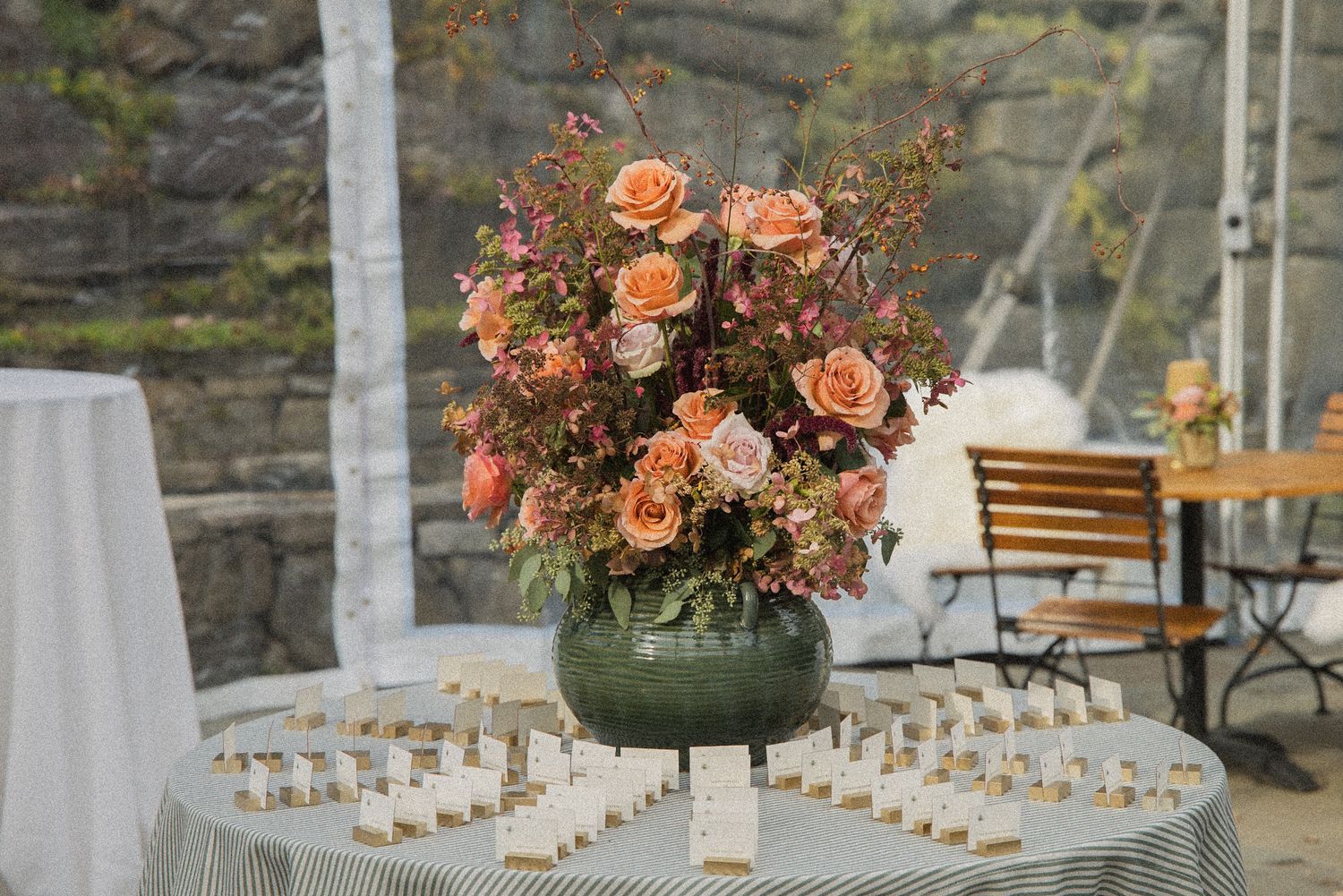 Peach and pink rose arrangement in a green vase sits on a striped table beside rows of wedding place cards.