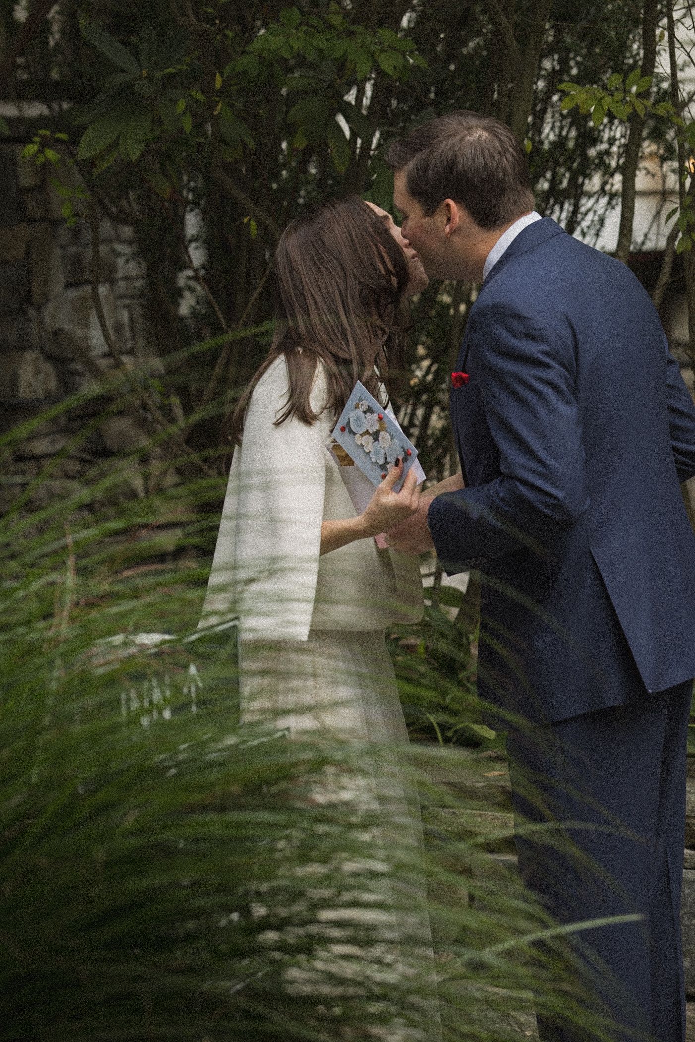 A bride in a white jacket and gown holds a floral card while kissing her groom, who wears a navy suit, beside tall grasses.