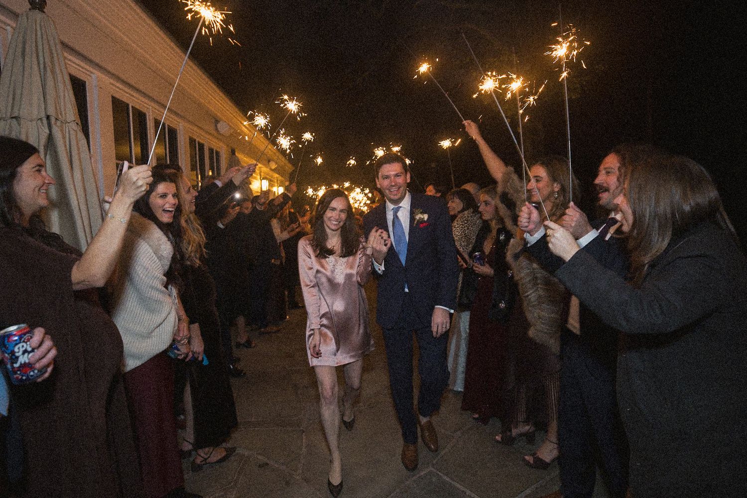 Smiling bride and groom walk hand in hand through a cheering crowd holding lit sparklers during a nighttime wedding exit.