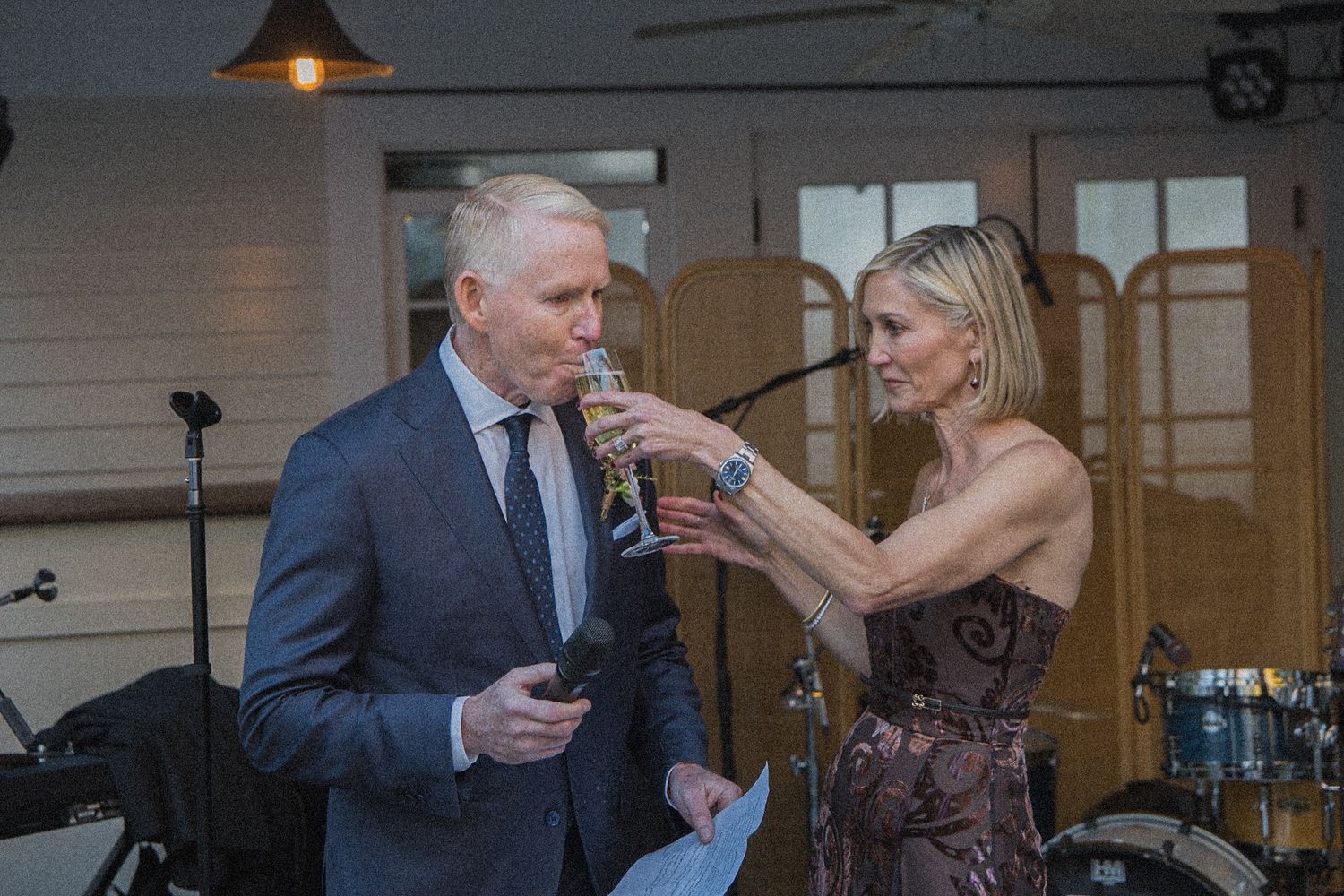 A man in a gray suit holding a microphone and paper toasts with a woman in a patterned dress raising a champagne flute at an event.