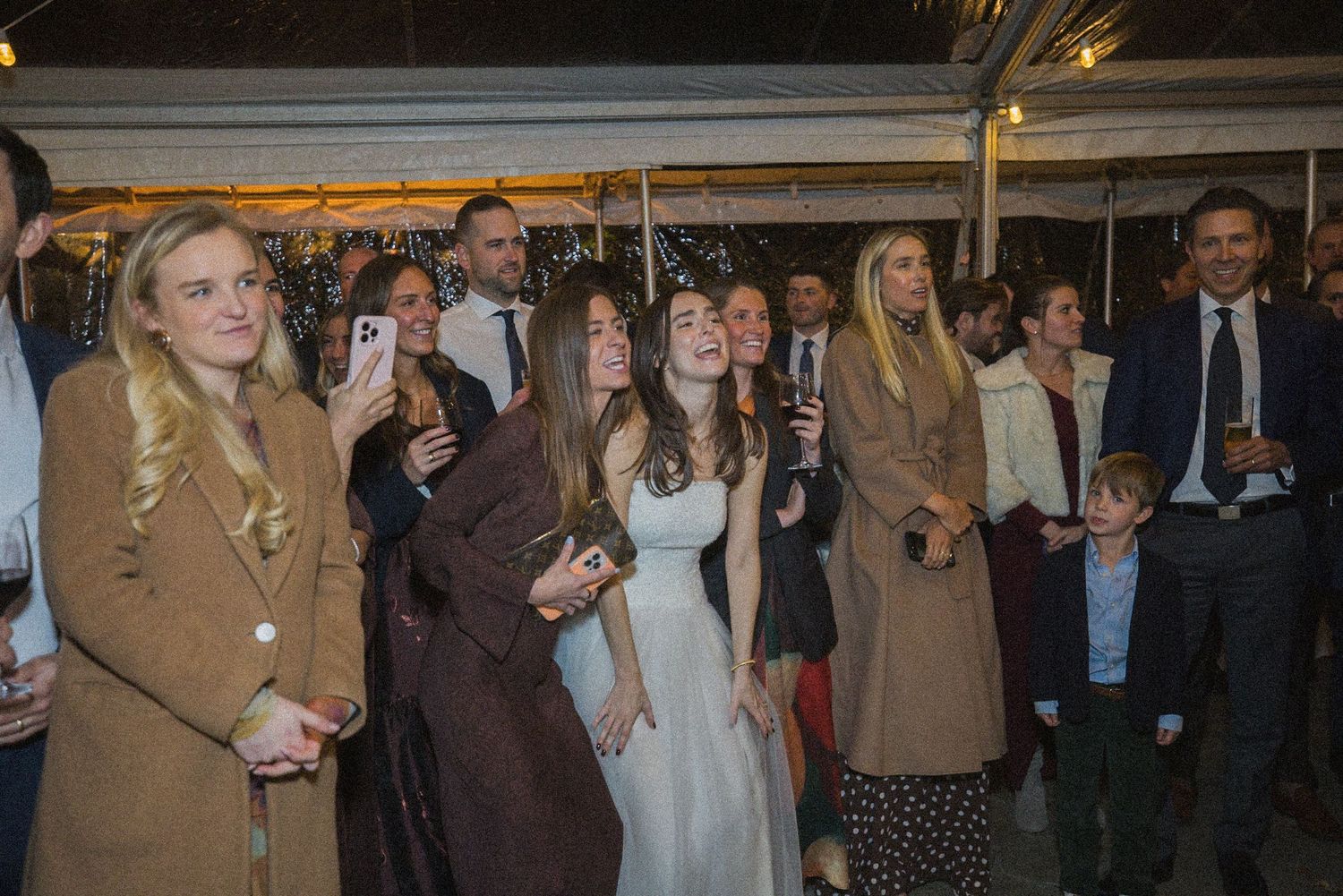 Smiling wedding guests in elegant evening attire watch and film the festivities under a twinkle-lit tent.