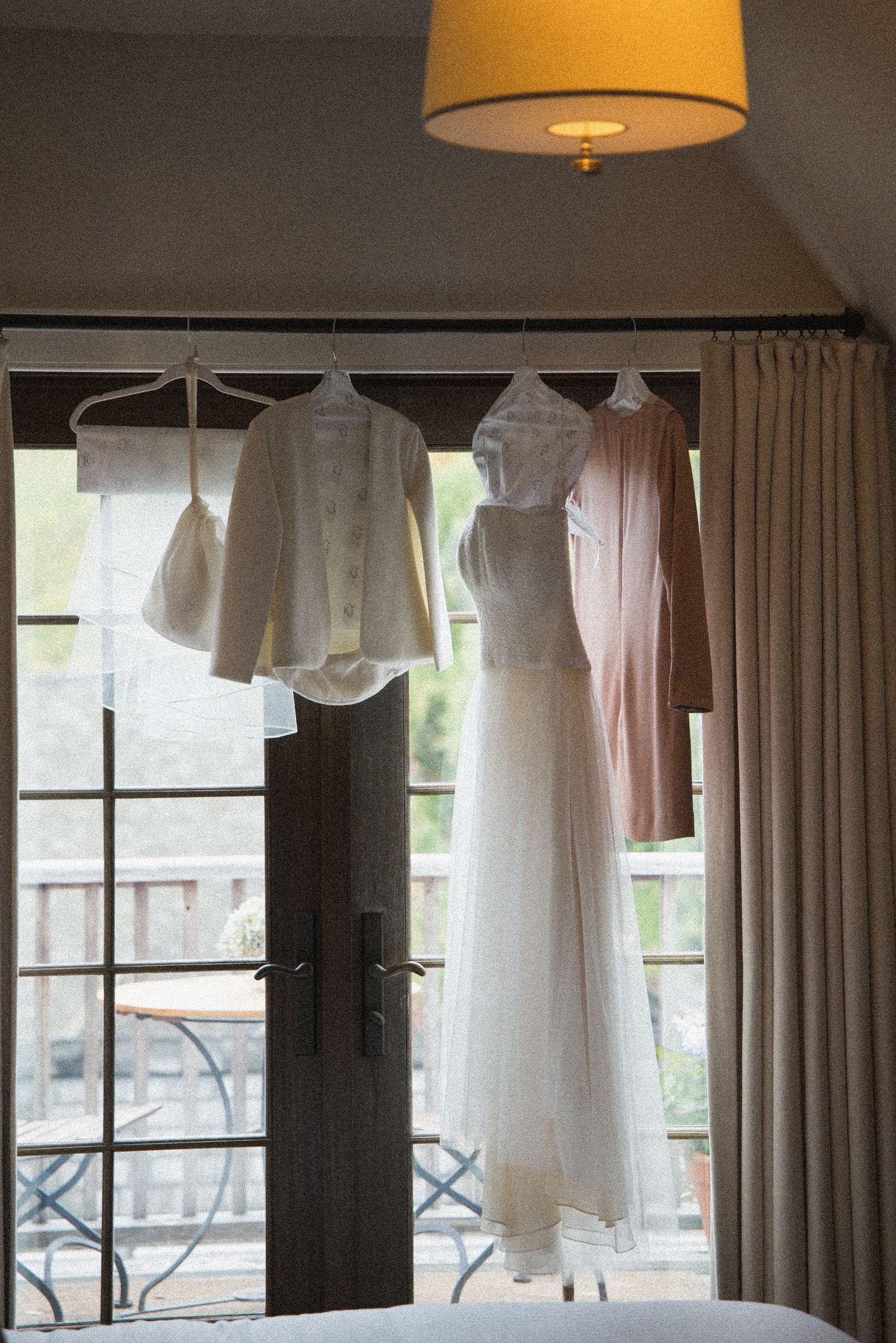 White wedding dress and bridal jackets hanging on hangers in front of French doors with a balcony view, beside a warm pendant lamp.