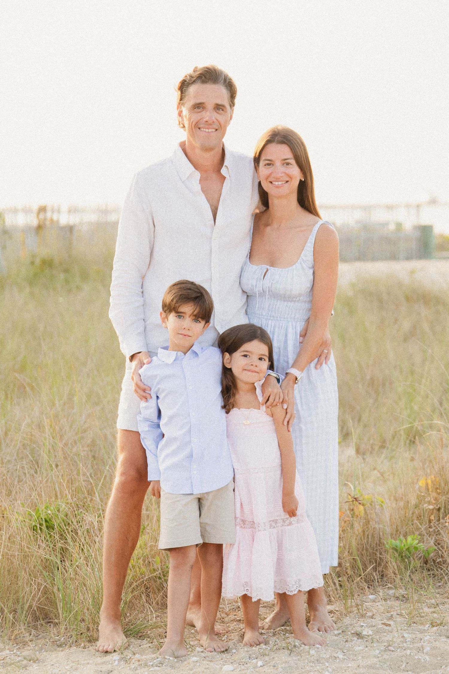 A happy family of four poses together on a sandy beach path, dressed in light summer clothing.