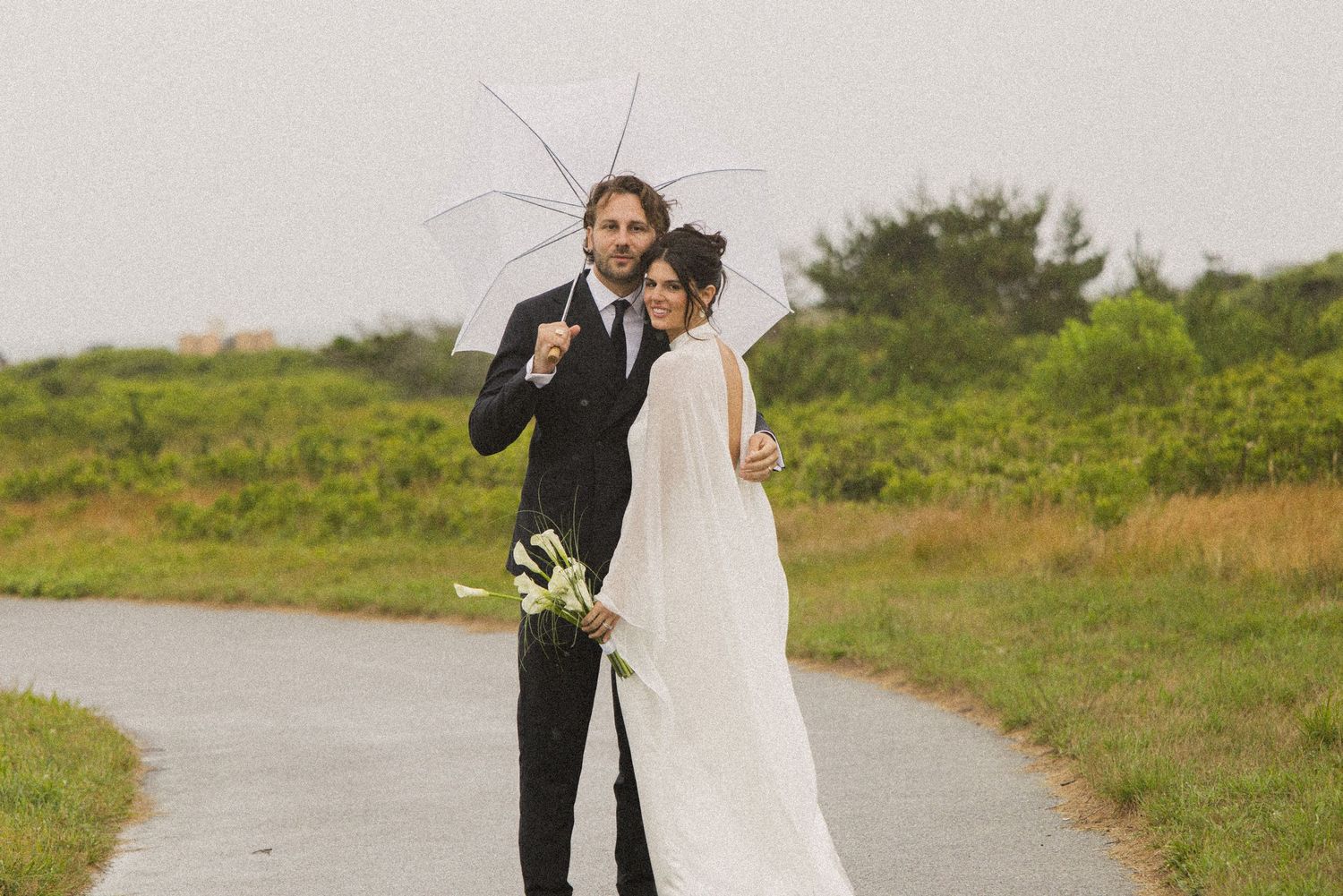 Bride and groom share umbrella on rainy wedding day, posing on path with lush green landscape backdrop.