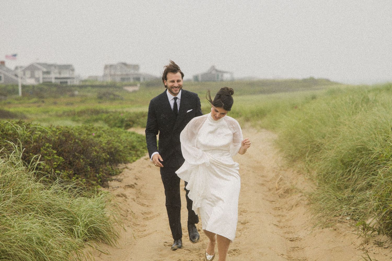 Couple in wedding attire walking hand-in-hand on a sandy coastal path surrounded by green grass on a misty day.