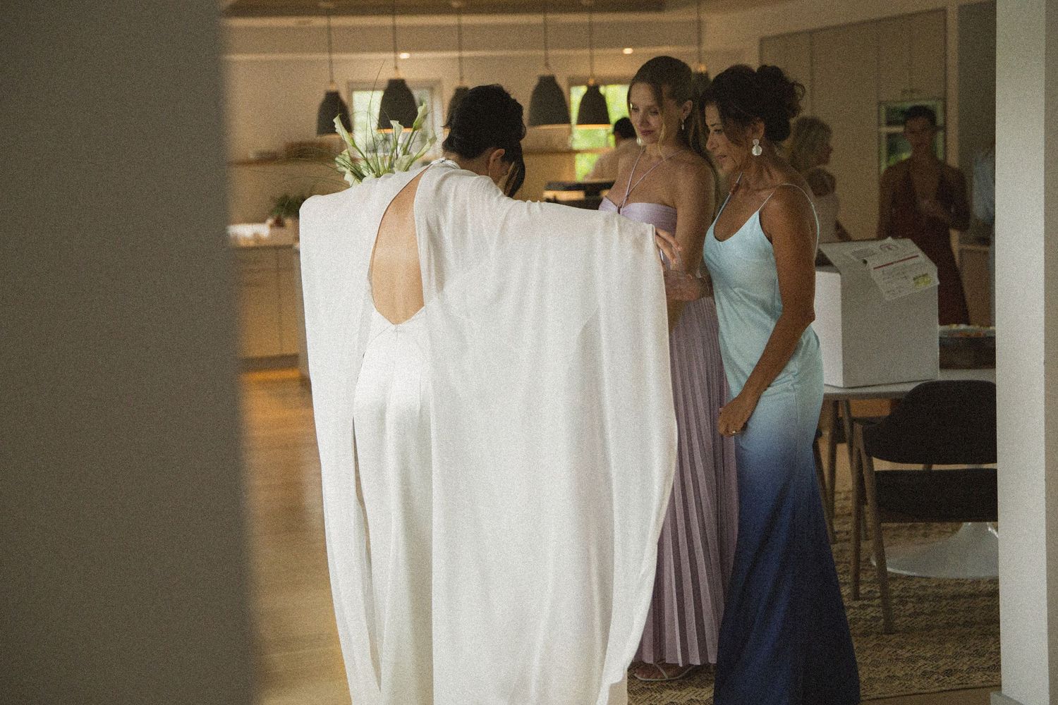 Bride in white gown with bridesmaids in purple and blue dresses preparing together indoors.