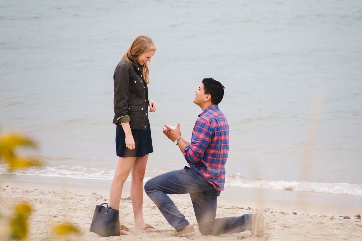 Man proposing to woman on a sandy beach, kneeling on one knee while presenting a ring box outdoors.