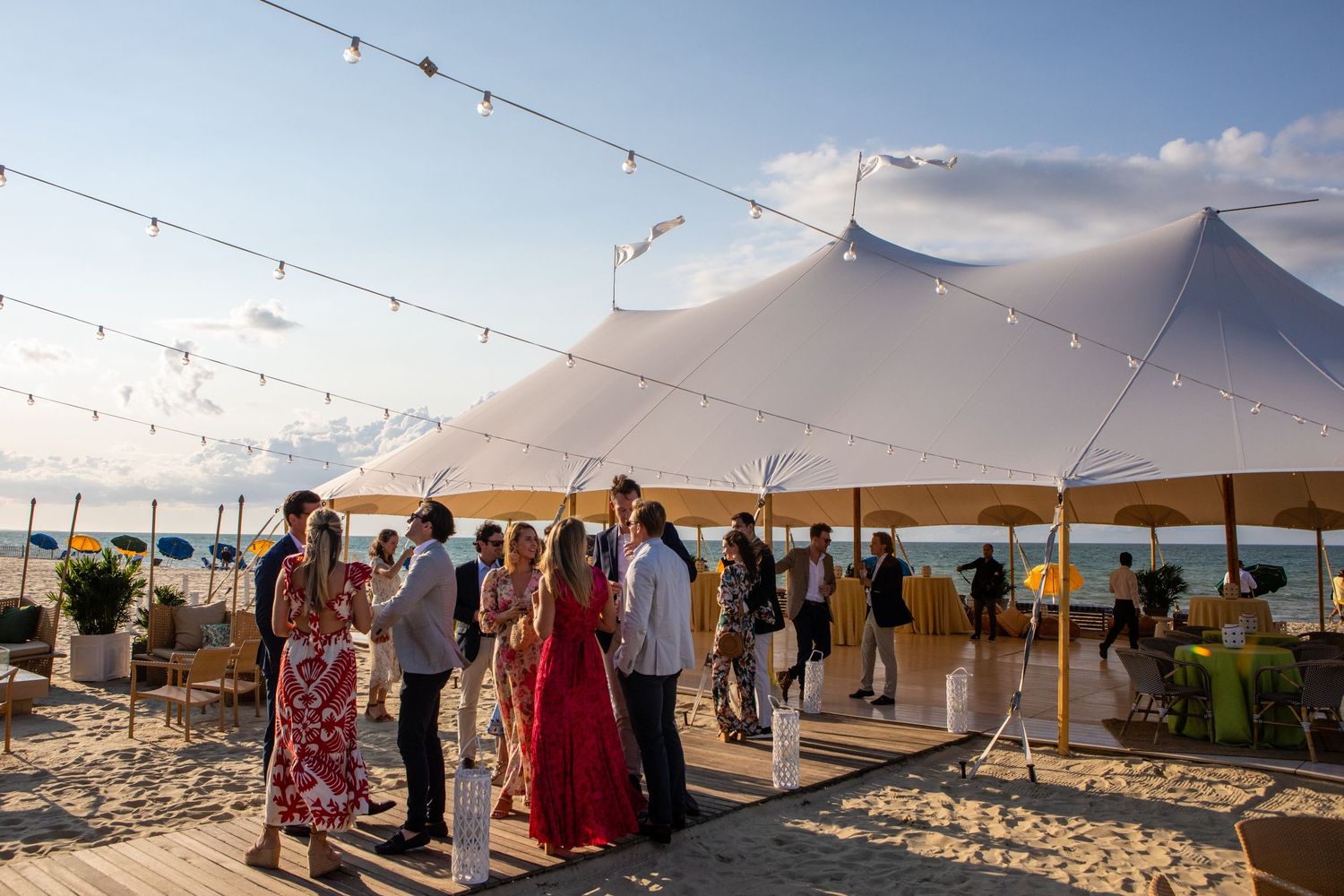 Guests mingle at an outdoor beach wedding reception under a white tent with string lights at sunset.