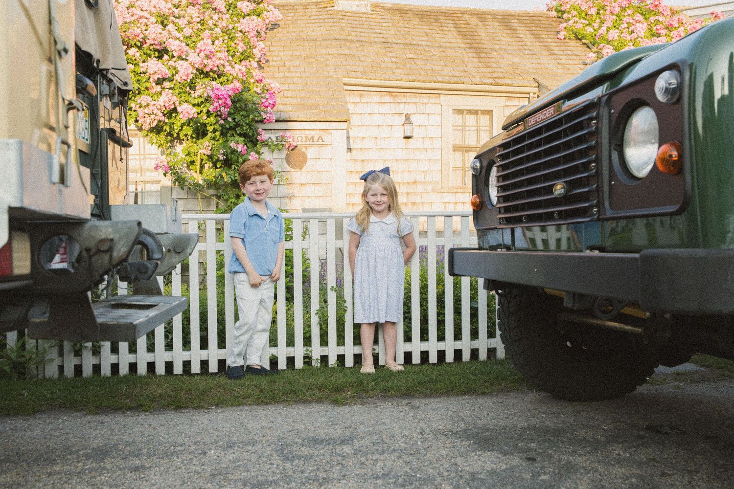 Two children stand between vintage vehicles by a white picket fence, with blooming pink flowers and a cottage in the background.