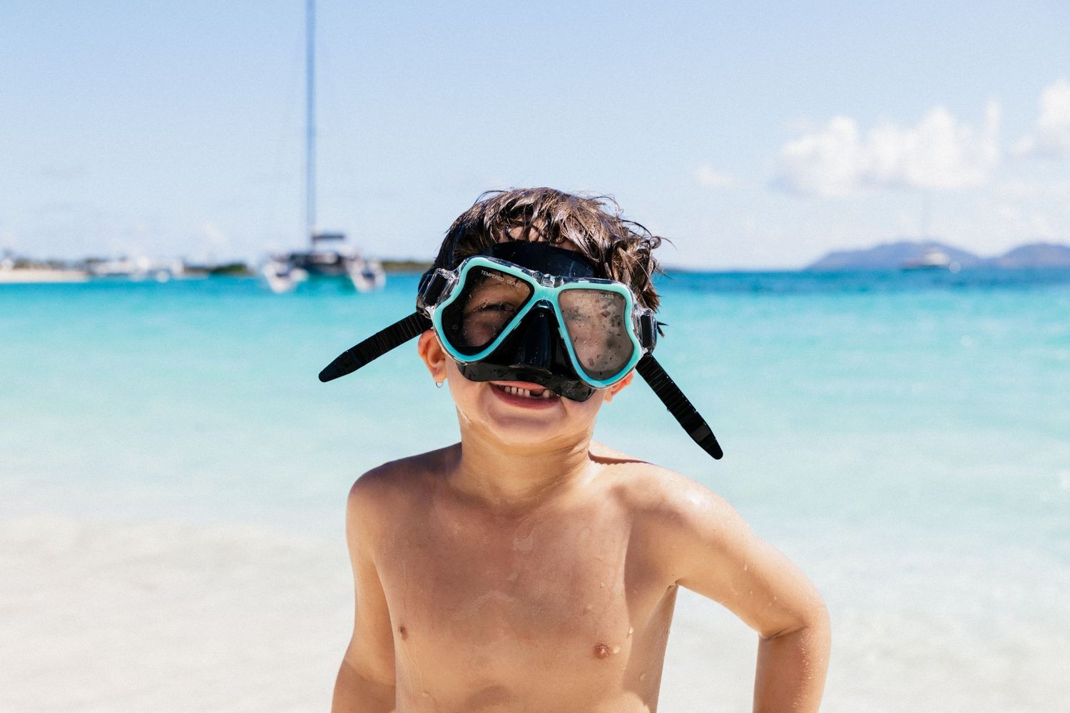 Young beachgoer wearing oversized snorkel goggles smiles on a tropical beach with turquoise water.