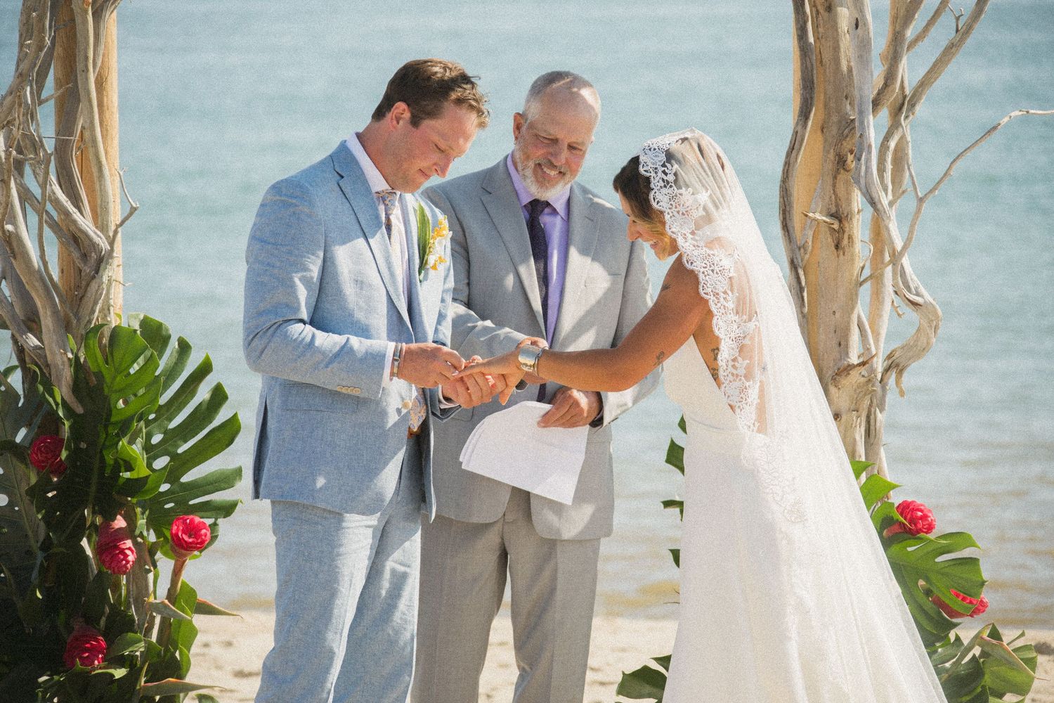 Wedding ceremony under driftwood arch decorated with red flowers on sandy beach.