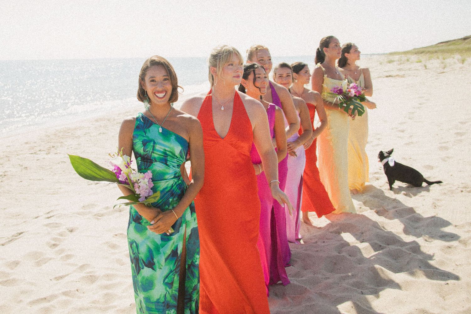 Bridesmaids in colorful dresses walk along sunny beach during wedding celebration.
