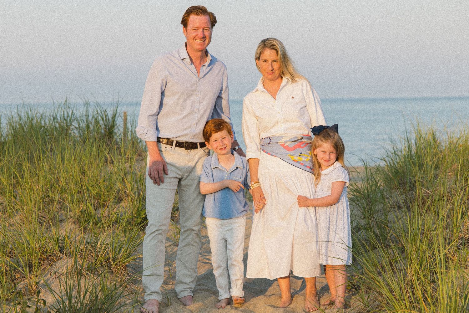 Family of four posing on sandy beach dunes with ocean backdrop during golden hour sunset.