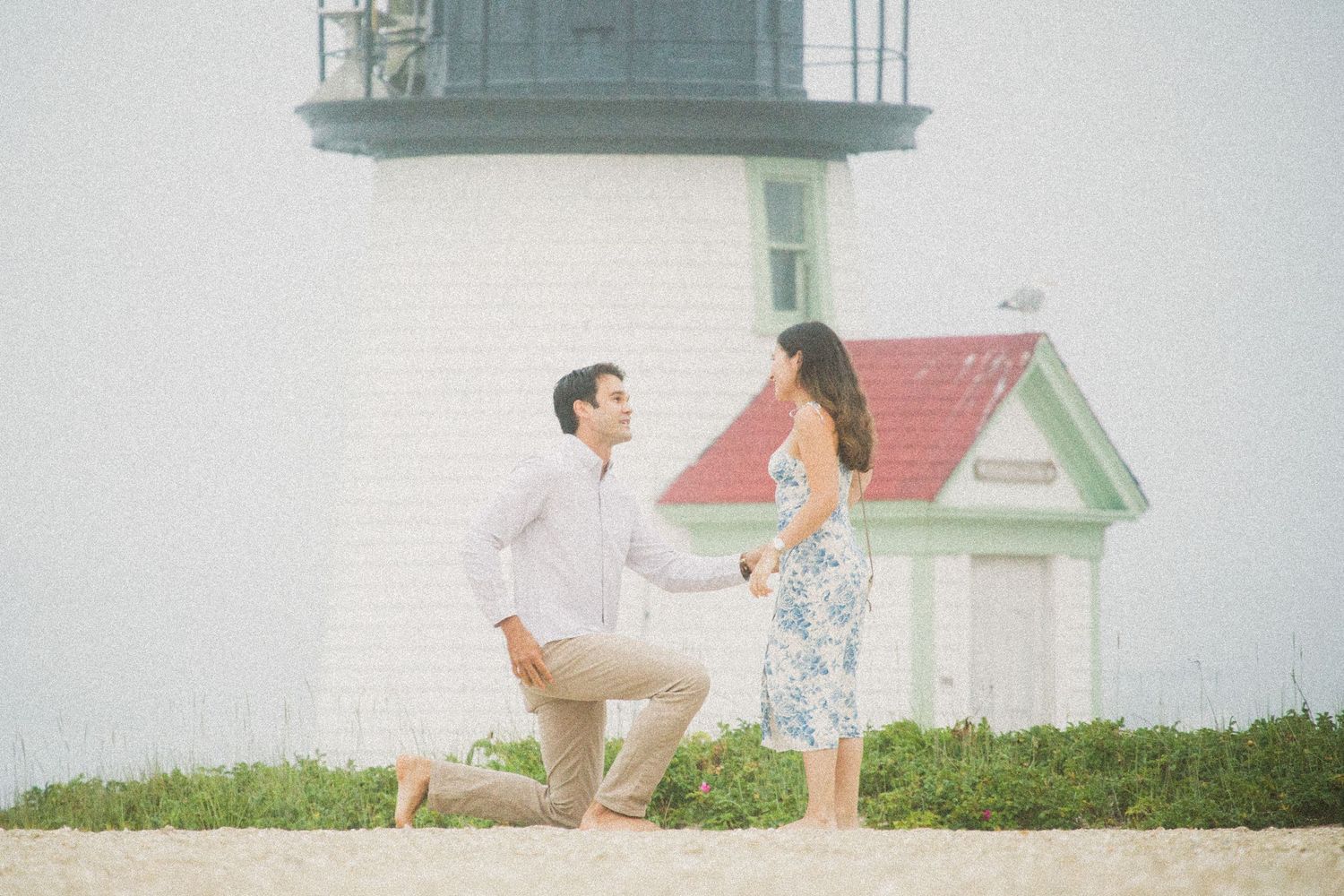 Man proposing on one knee to woman near a lighthouse on a sandy beach.