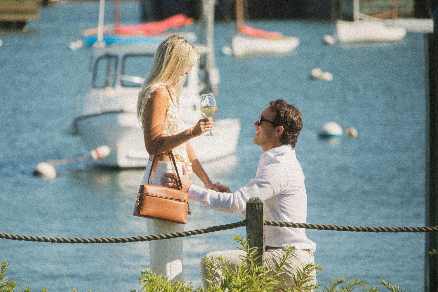 Two people sit together on a dock overlooking a marina with boats and blue water on a sunny summer day.
