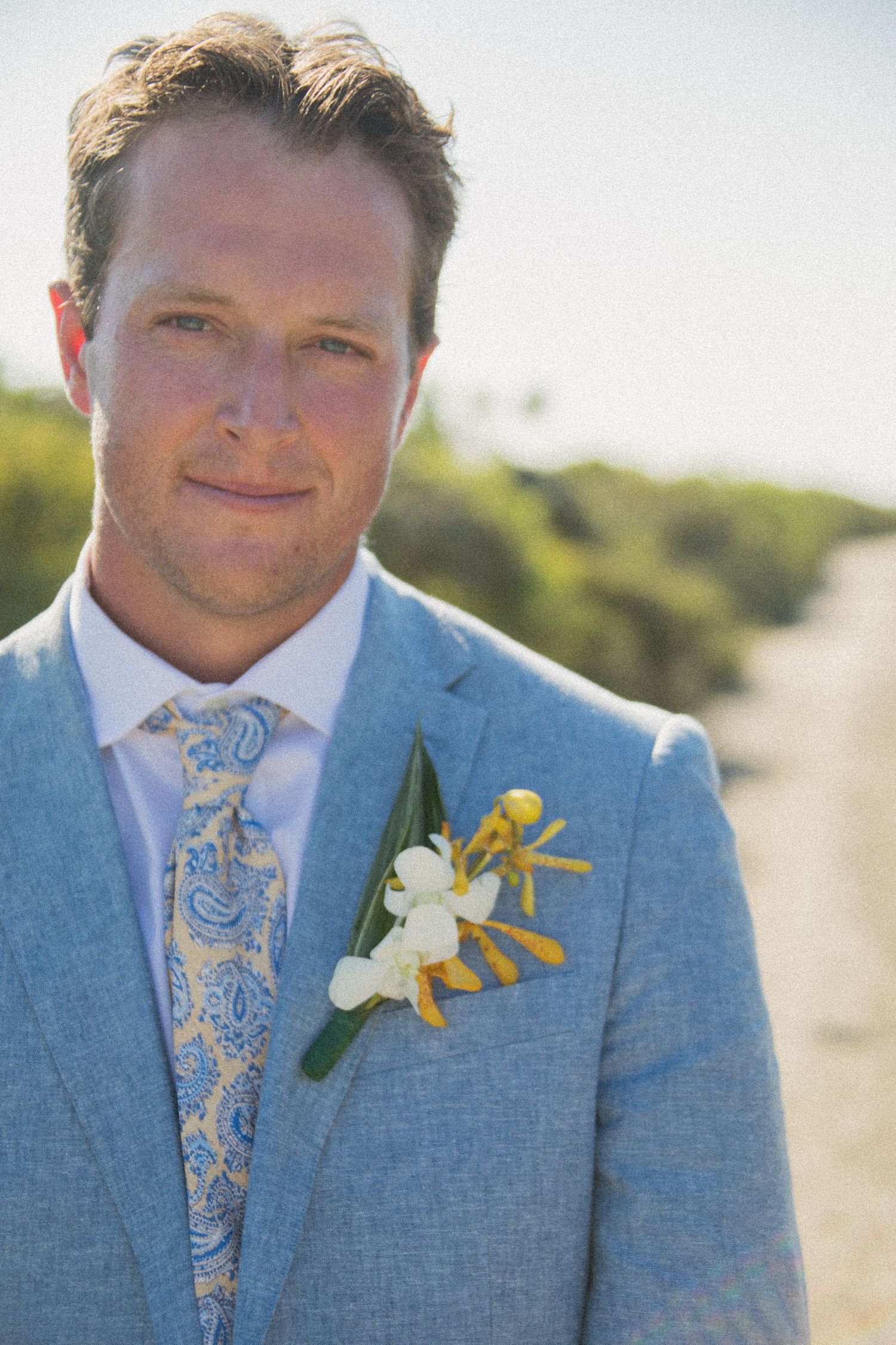 Groom wearing light blue suit with floral tie and boutonniere poses outdoors against natural backdrop.