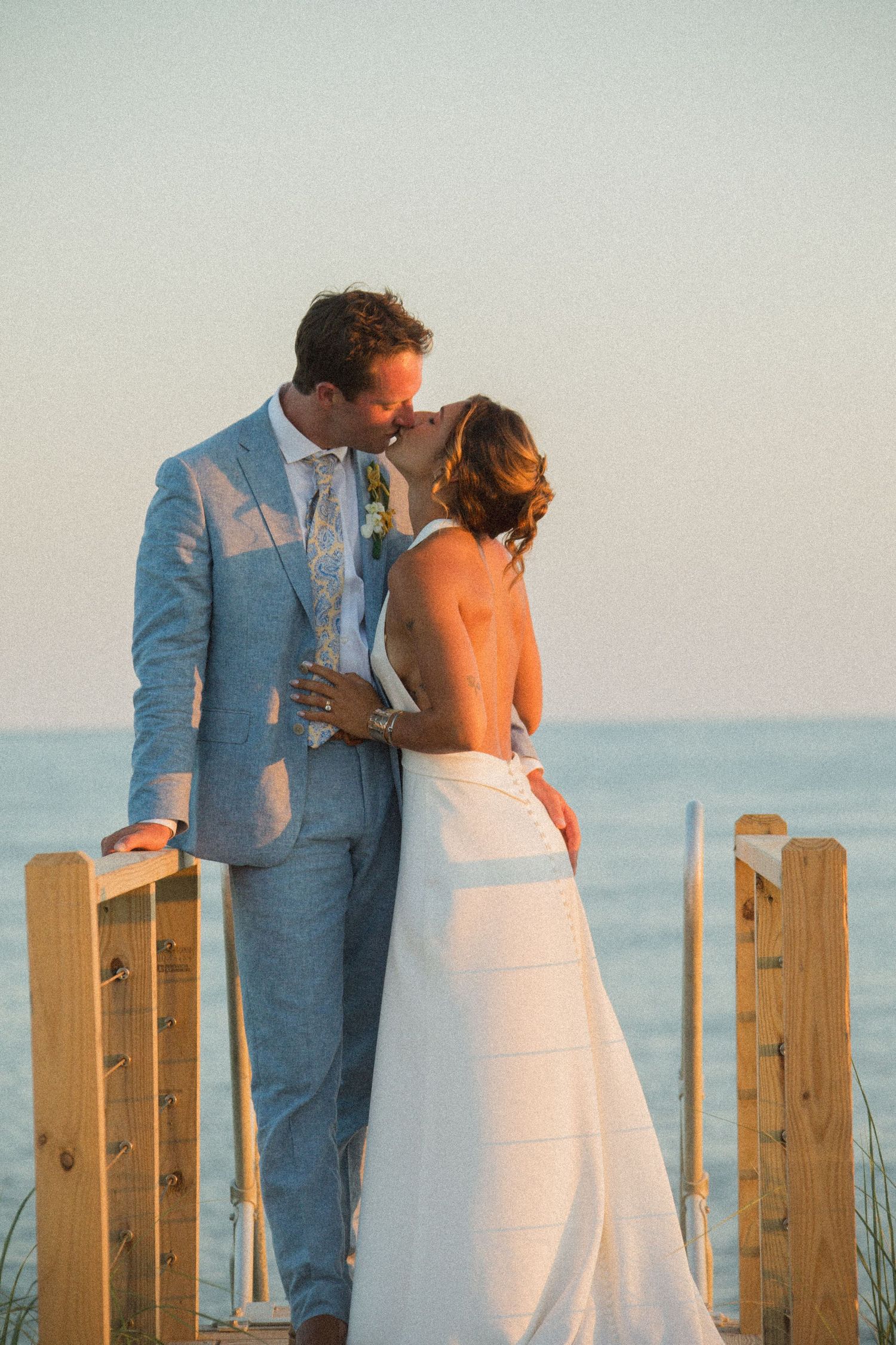 A couple in blue suit and white dress share a tender moment by a wooden fence near the ocean.