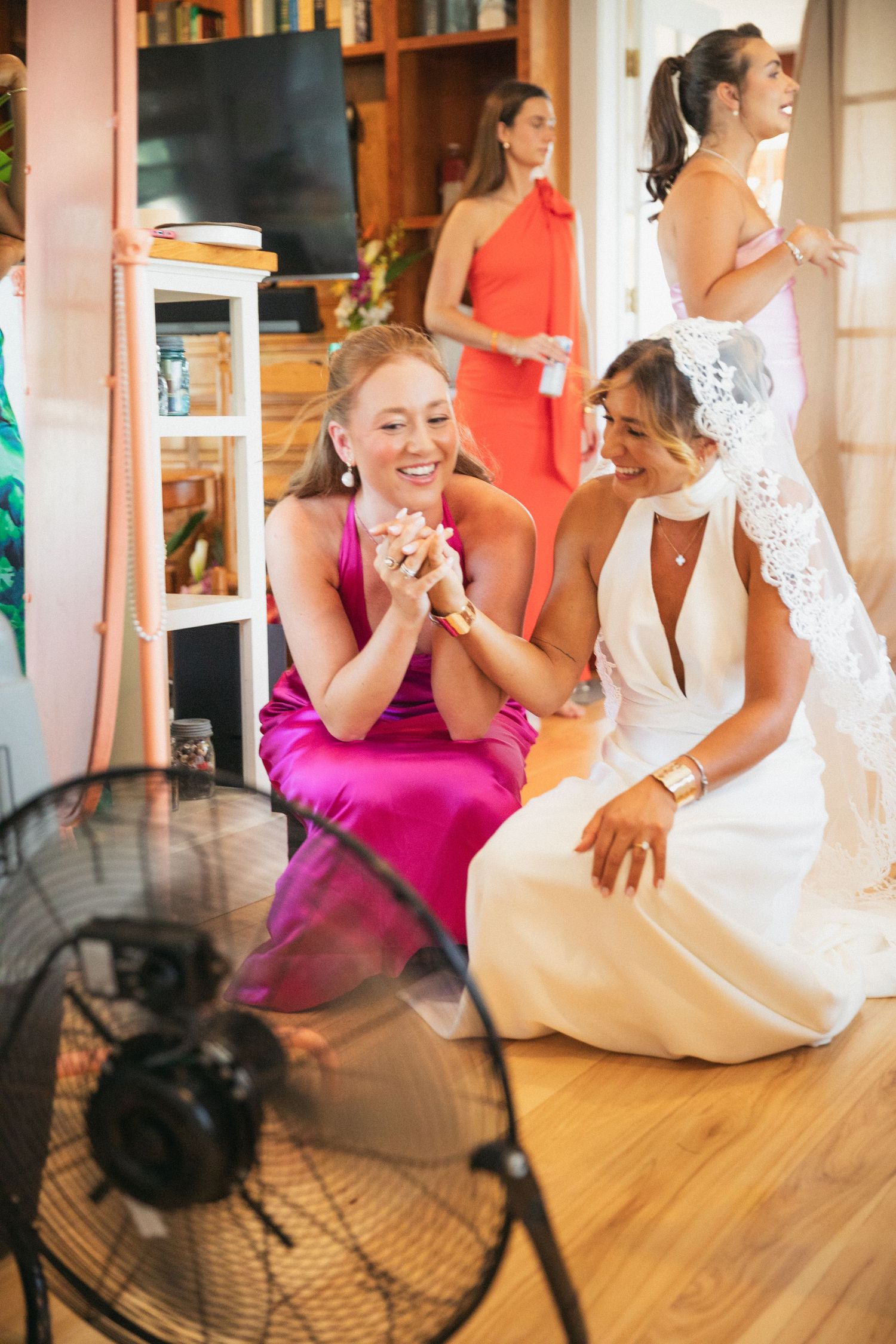 Wedding party gathering in a living room with a floor fan and colorful dresses.
