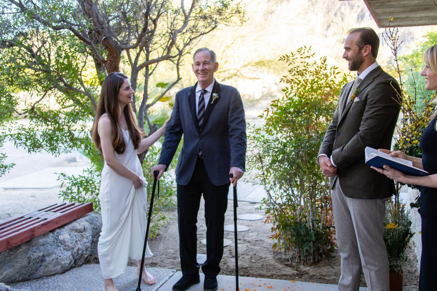 A couple exchanges vows in front of a white brick wall decorated with yellow flowers and branches.