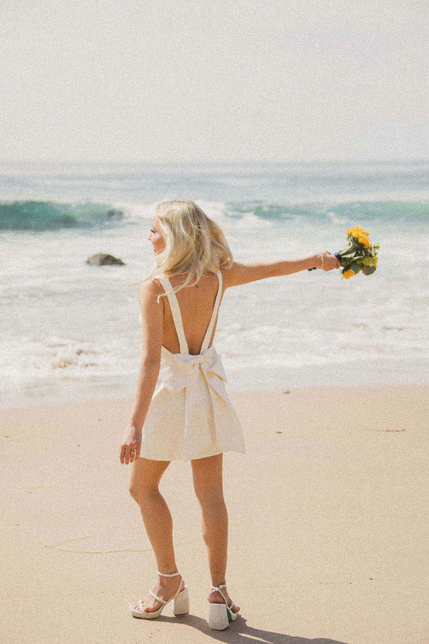 Bride dances on beach in short white dress while holding yellow sunflower bouquet.