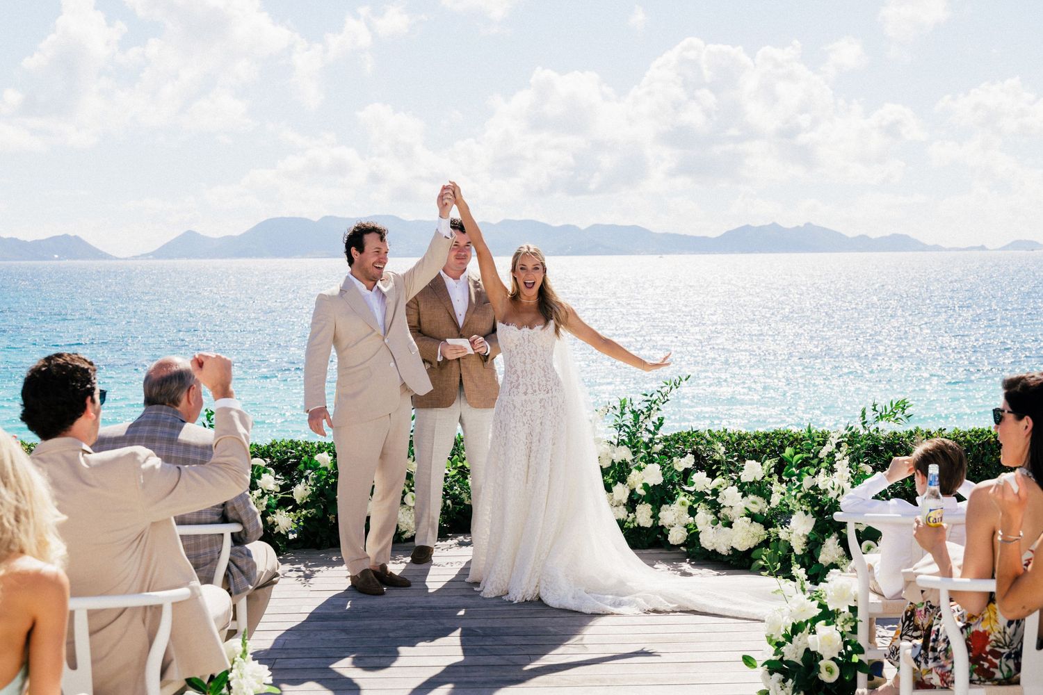 Wedding guests celebrate on a scenic white terrace overlooking a beautiful ocean view with mountains in the distance.