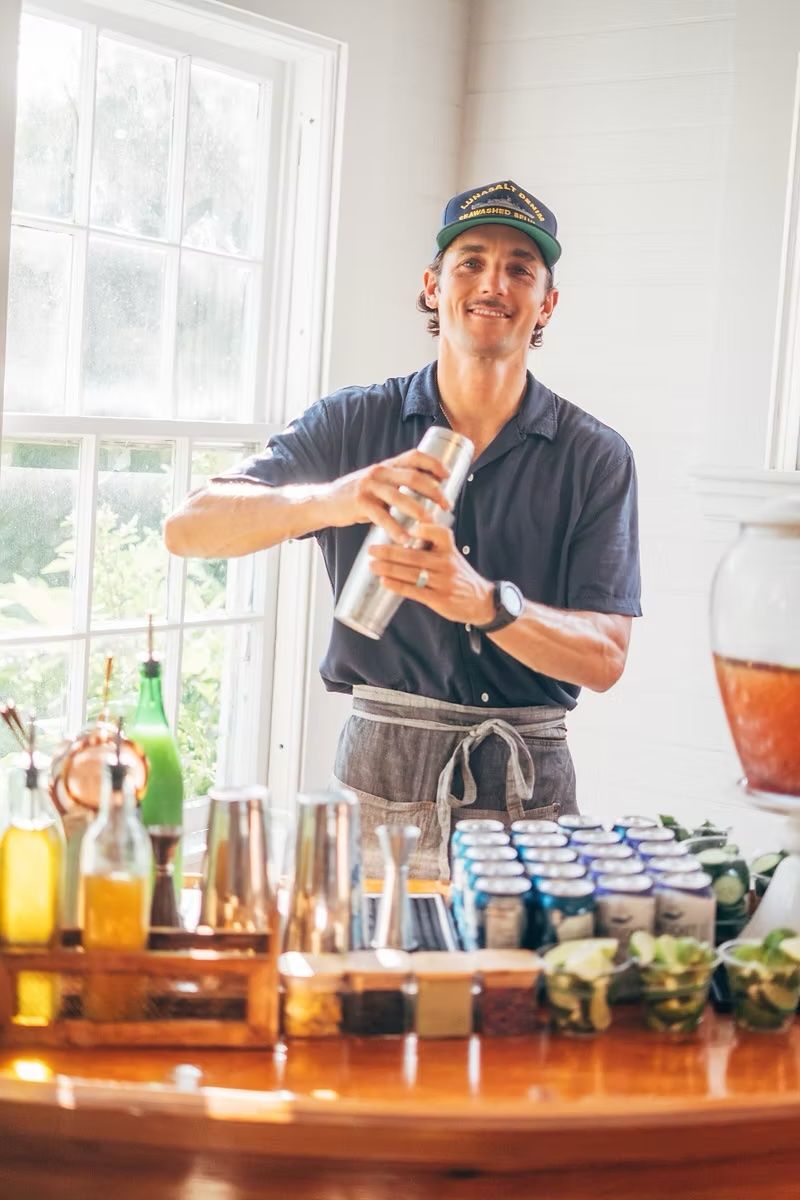 Smiling bartender in cap and apron shaking cocktail shaker at bar setup with bottles and glasses.