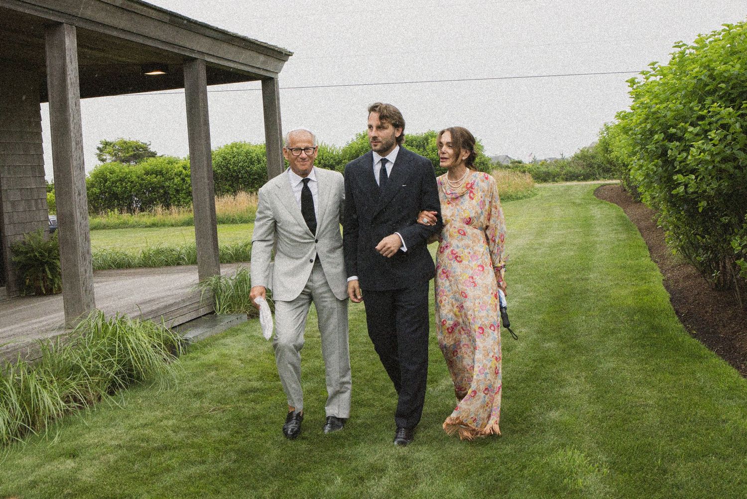Three elegantly dressed people walking on a green lawn outside a venue, wearing formal attire including suits and a floral gown.