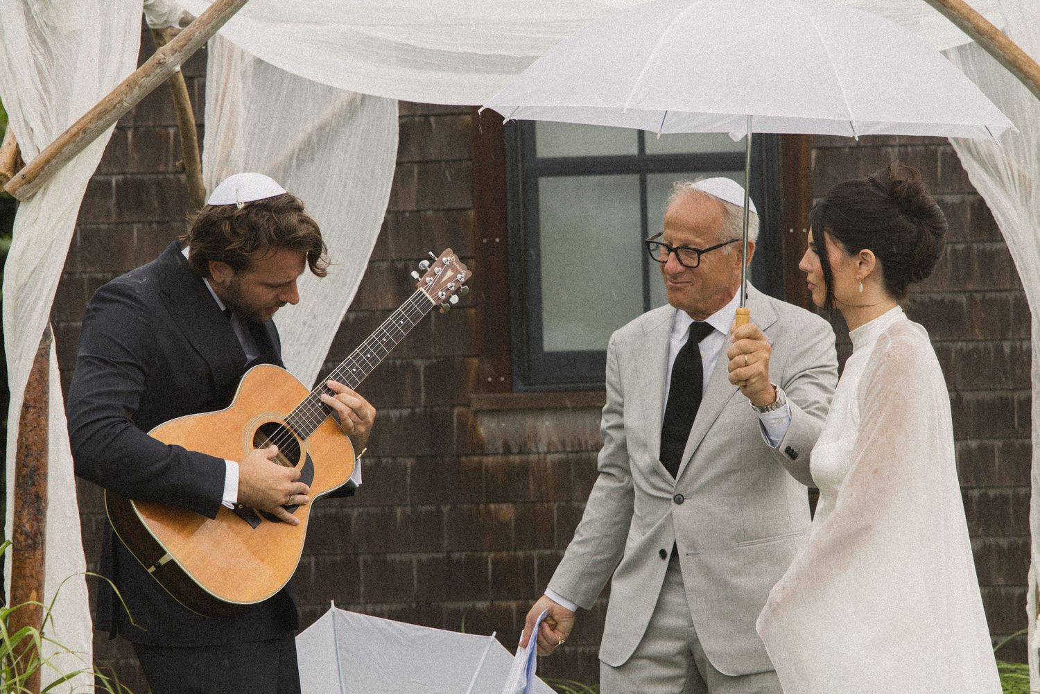 Guitarist plays at outdoor Jewish wedding ceremony under chuppah as officiant raises glass with bride.