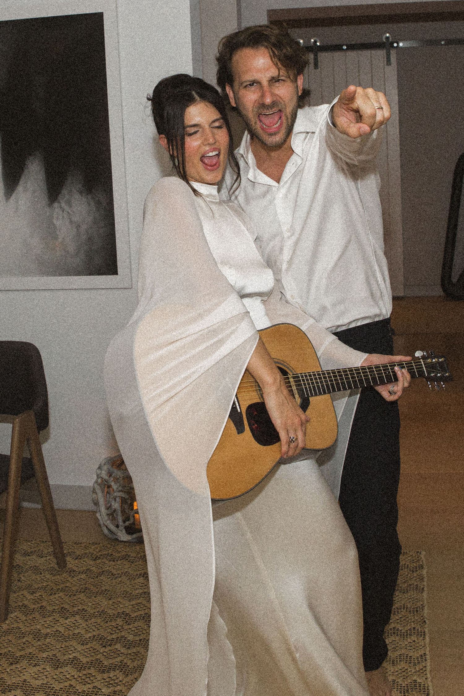 Two people in white outfits pose with an acoustic guitar, smiling and pointing in a bright indoor setting.
