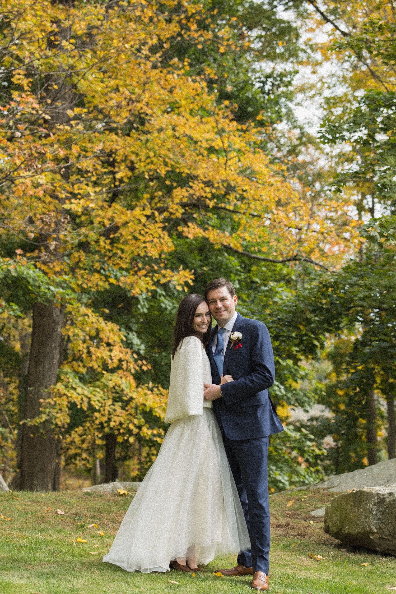 Bride in a white tulle gown and cream jacket embraces a groom in a navy suit on a lawn beneath golden autumn trees.