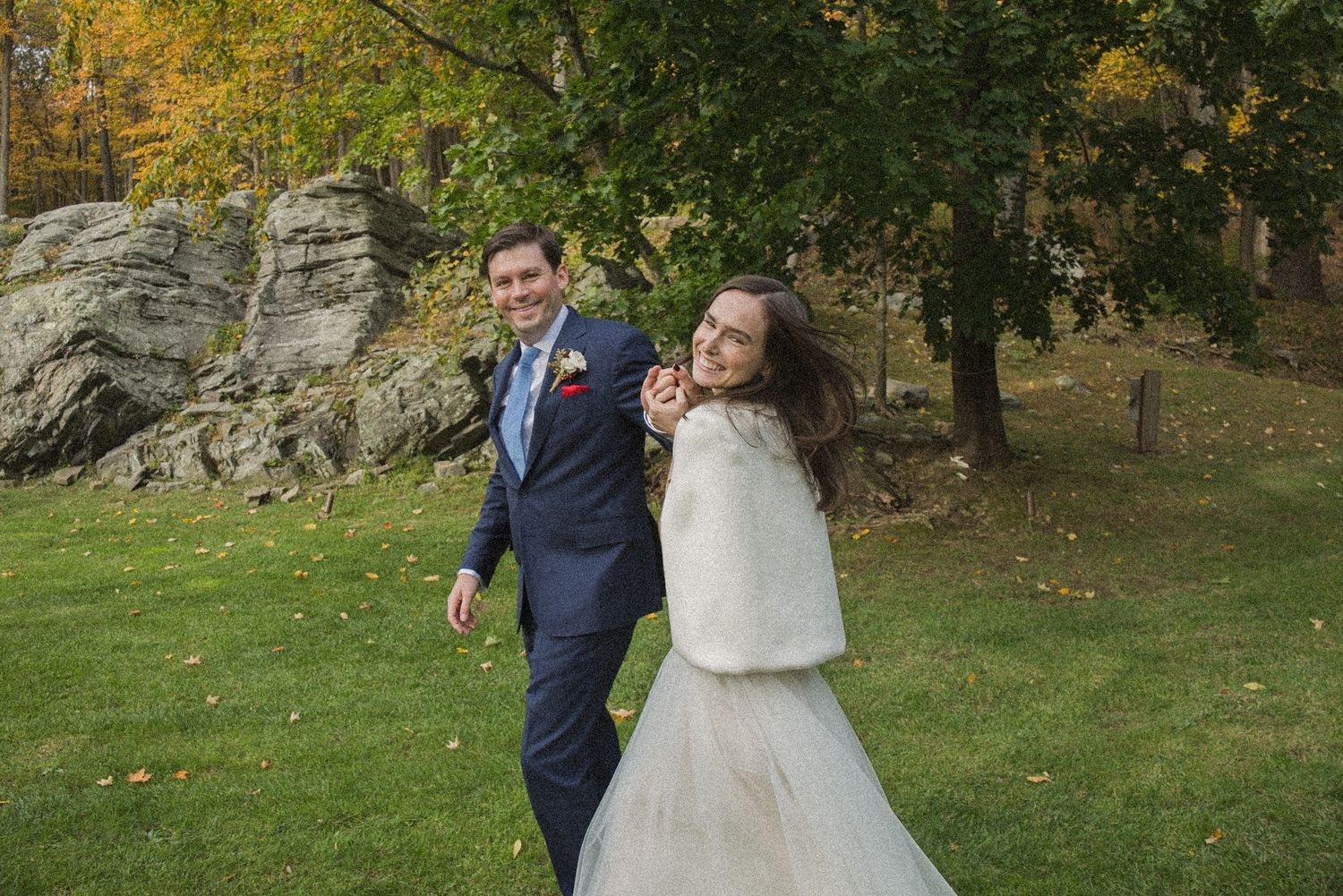 Smiling bride in white gown and fur shawl holds hands with her groom in a navy suit while walking across a lawn near rocks and fall foliage.