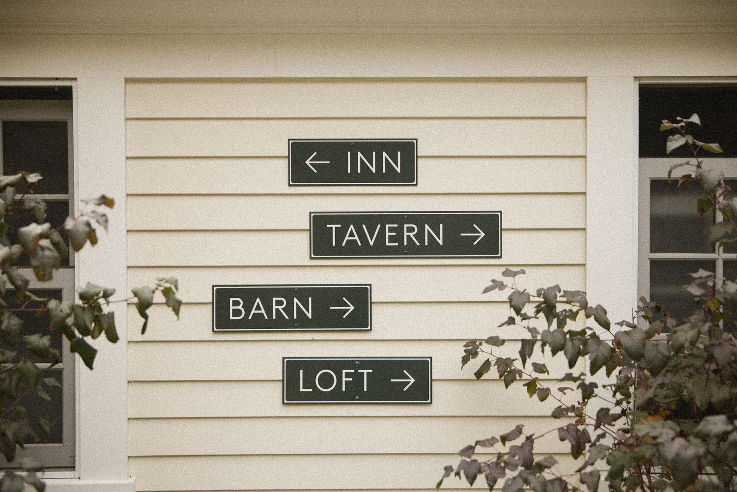 Four dark green directional signs reading Inn, Tavern, Barn and Loft with arrows, mounted on cream clapboard siding beside leafy branches.