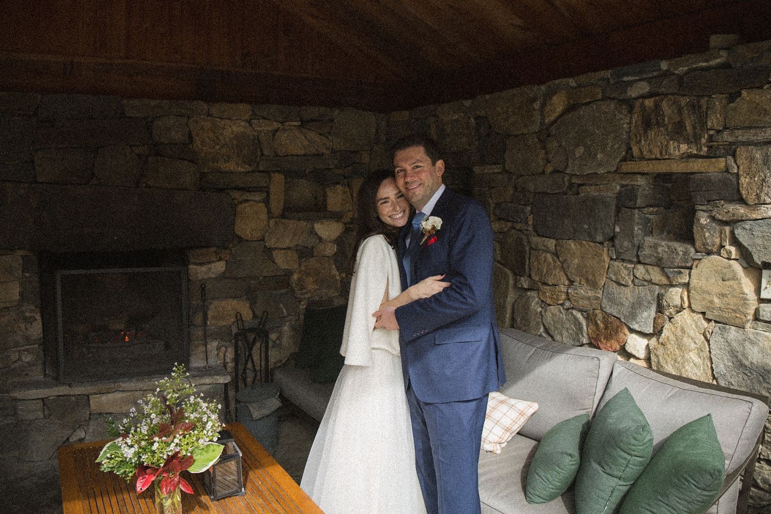 A bride in a white gown and a groom in a navy suit embrace beside a rustic stone fireplace and a cozy sofa with green pillows.