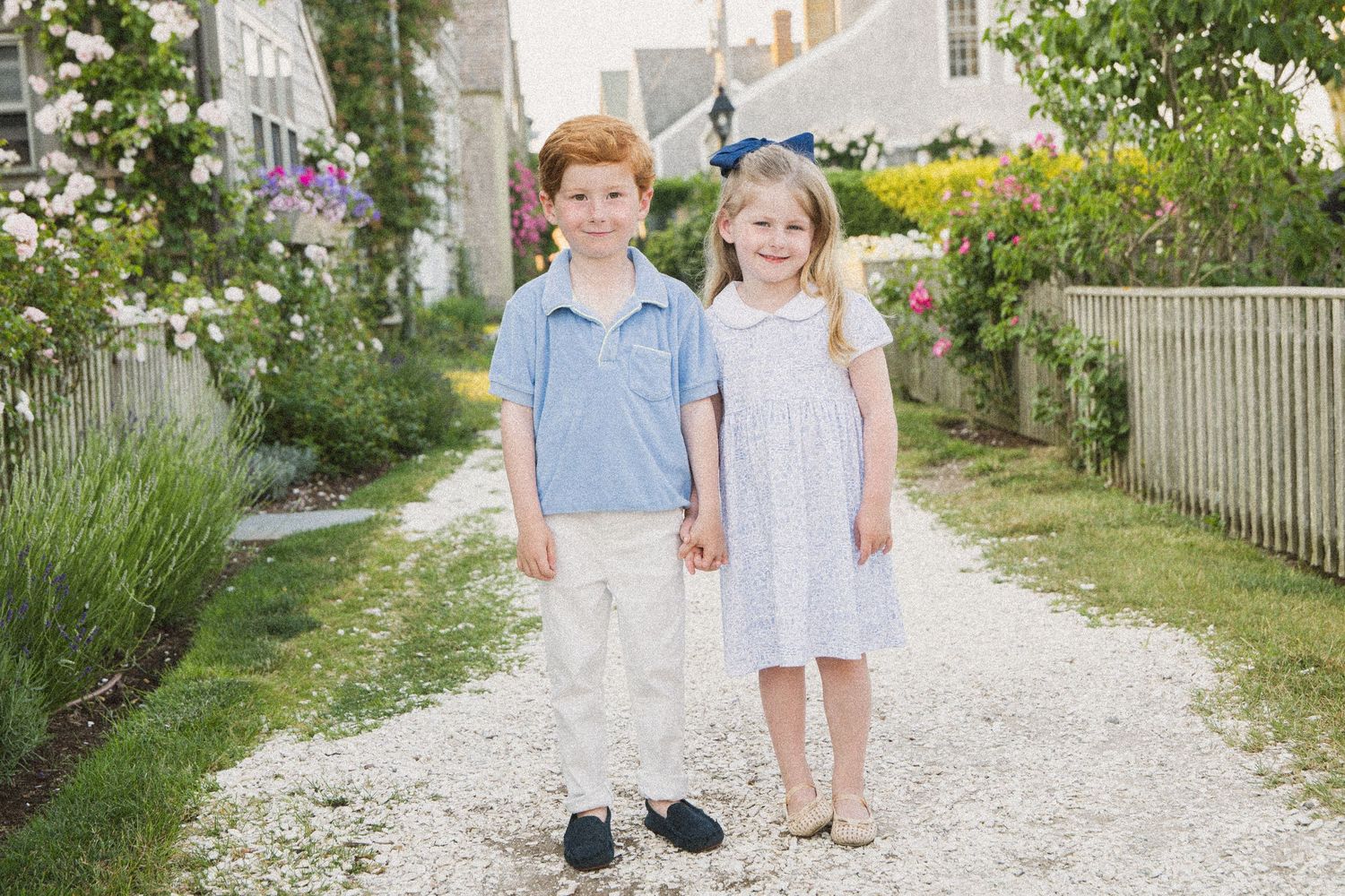 Two young children, a red-haired boy and a blonde girl, hold hands on a garden path surrounded by flowers.