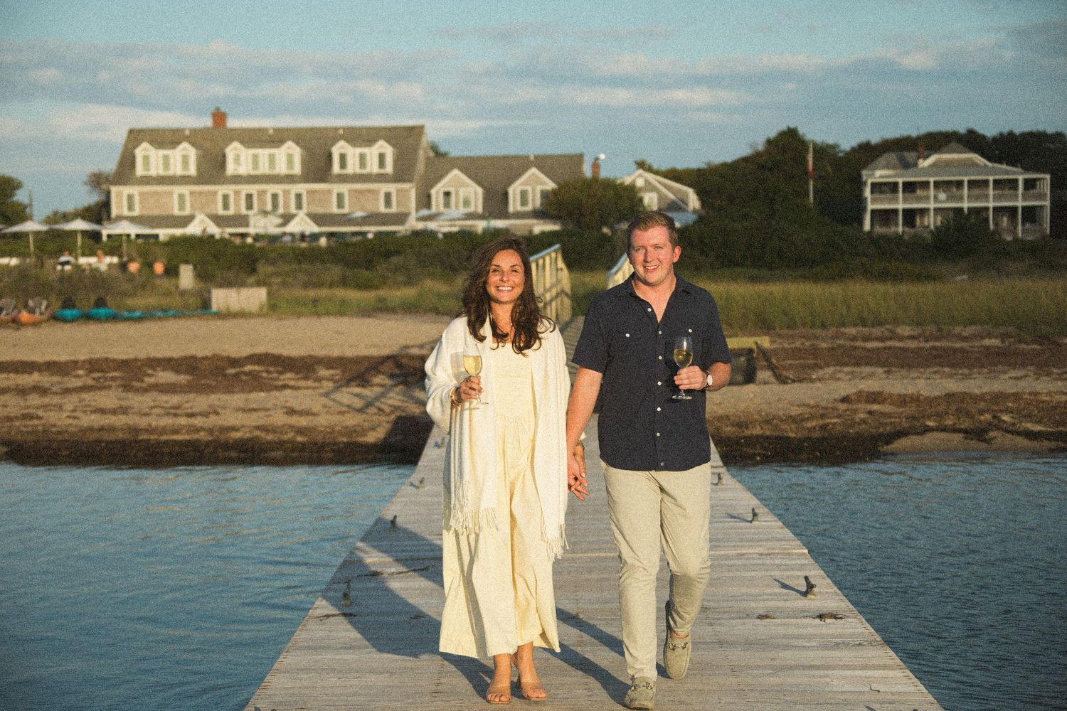 A couple walks along a coastal boardwalk with beach houses in the background.